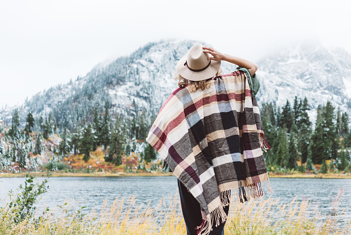 Styish woman feeling happy among amazing mountains, enjoy the nature landsape. Forest and lake, wearing hat and poncho, boho and wanderlust style