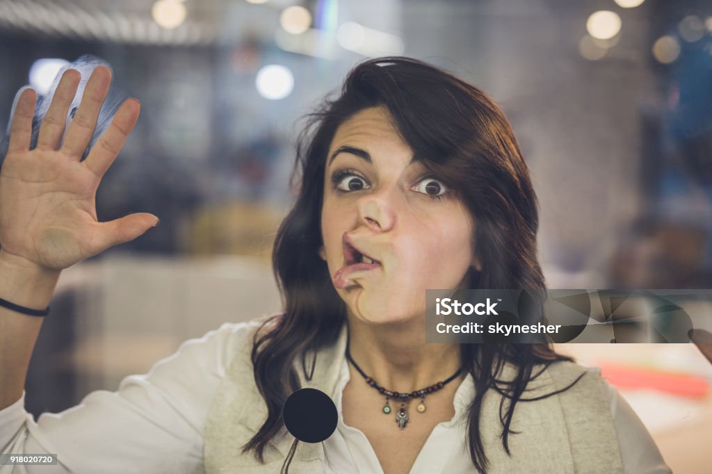 Playful woman with glued face on glass wall looking at camera. Young freelance worker sticking her face on a glass wall and looking at camera. The view is through glass. Human Face Stock Photo