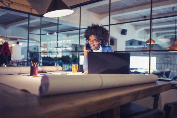 Working Late. Young woman, architect working late in office Young African-American woman architect working on a project. She is working late after work. She is talking on mobile phone, using laptop african american architecture stock pictures, royalty-free photos & images