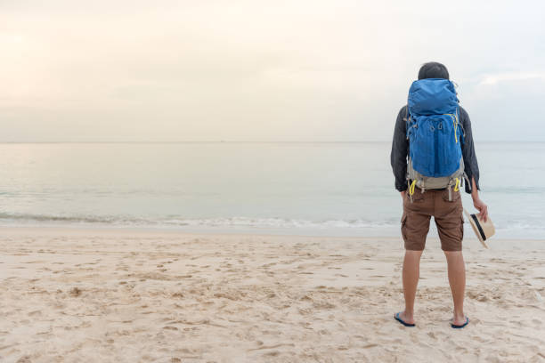 istock Young Asian backpacker man standing on the beach and holding hat 918042152