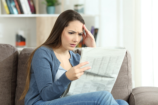 Portrait of a concerned woman reading bad news in a newspaper sitting on a couch in the living room at home