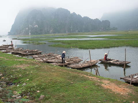 Ninh Binh (UNESCO World Heritage Site), VIETNAM – JANUARY 7, 2018: Adult Vietnamese woman in a straw conical hat sits in a wooden boat and rowing oars against the background of a mountain and misty sky in the province Ninh Binh, Vietnam