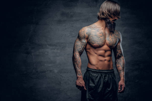 Hipster male with tattooed muscular body. Shirtless athletic bearded hipster male with tattooed muscular body on grey vignette background. cross shoulder tattoos stock pictures, royalty-free photos & images