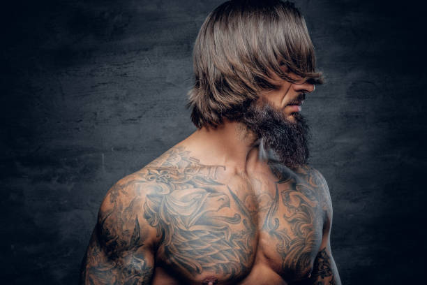 Bearded muscular brutal male on grey background. Muscular bearded male with tattooed body isolated with illumination on grey background. cross shoulder tattoos stock pictures, royalty-free photos & images