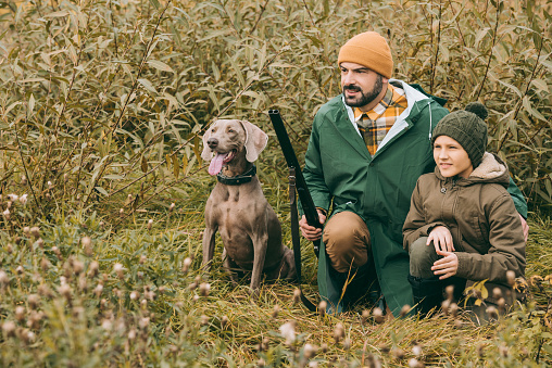 Father and son squatting in bushes with a dog and gun at hunt