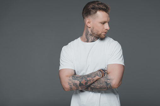 stylish young tattooed man in white t-shirt standing with crossed arms and looking away isolated on grey