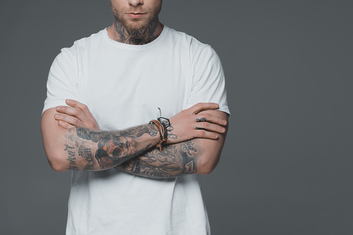 cropped shot of young man with tattoos standing with crossed arms isolated on grey