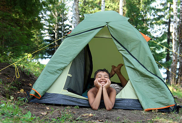 Young man wearing a beanie and headphones sitting in a grassy area, packing a small tripod into his backpack next to a camping tent, enjoying a break during a solo adventure