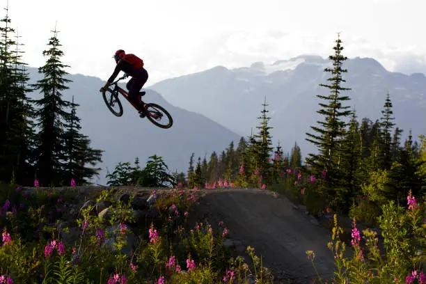 A male mountain bike rider hits a jump in the mountains of British Columbia, Canada. He is wearing a full face helmet and is riding an enduro-style mountain bike.