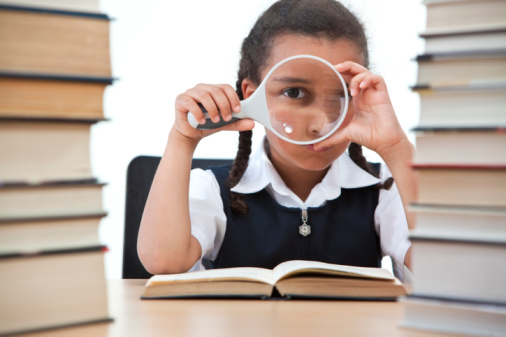 A beautiful young mixed race girl looking through a magnifying glass while reading in a school classroom with two piles of books in front of her.