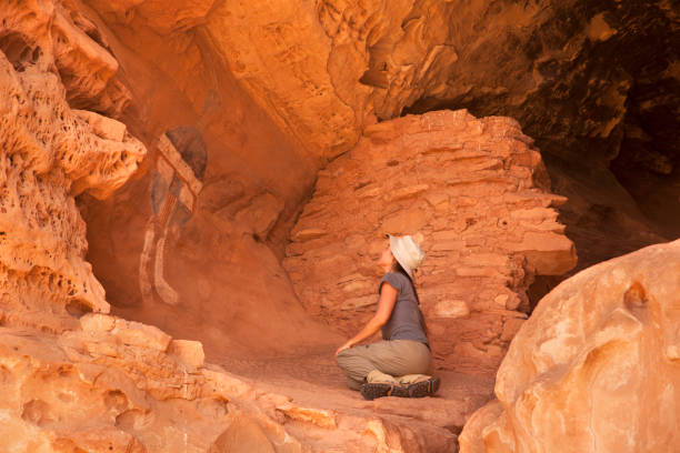 Woman hiker admires All American Man rock art Canyonlands National Park pueblo ruin Utah Sitting in a sandstone cave next to a ancient puebloan or Anasazi pueblo dating from A.D. 900 to 1300, a woman hiker admires the colorful red, white, and blue painted rock art with a shield pictograph known as the "All American Man" in Canyonlands National Park in Utah. canyonlands national park stock pictures, royalty-free photos & images