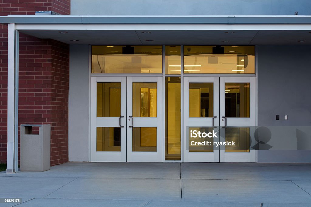 Entrance to a Modern Public School Building with Illuminated Interior Front entrance of a modern school building with metal and glass doors photographed at at dusk in the Northeastern USA using a Canon 5D MarkII DSLR. Interior is lit up and reflecting a subtle glow onto the concrete ground. School Building Stock Photo