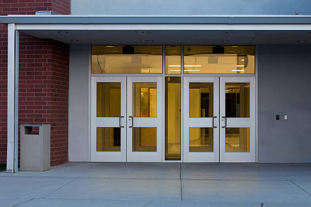 Entrance to a Modern Public School Building with Illuminated Interior Front entrance of a modern school building with metal and glass doors photographed at at dusk in the Northeastern USA using a Canon 5D MarkII DSLR. Interior is lit up and reflecting a subtle glow onto the concrete ground. Entrance Door stock pictures, royalty-free photos & images