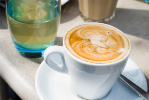 A cup of cappuccino, together with a blue glass of cold apple juice and a latte on a table outside.