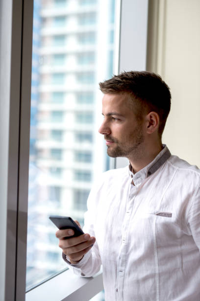 istock Man standing next to bright window holding phone, looking away. 918308312