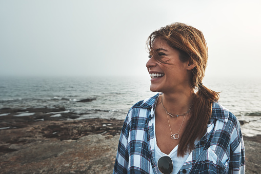 Shot of a cheerful young woman standing on rocks next to the ocean while laughing outside during the day