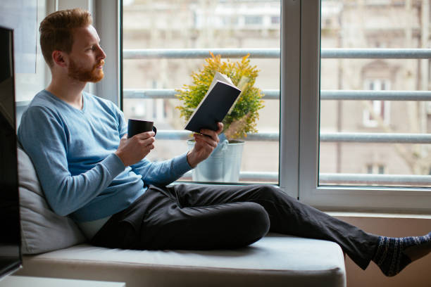 istock Man reading book at sofa near the window 918312268