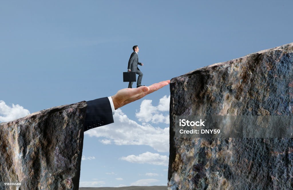Businessman Walking On Hand That Bridges The Gap A businessman holds his briefcase as he walks across a large hand that bridges the gap between two cliffs. Bridging The Gap Stock Photo
