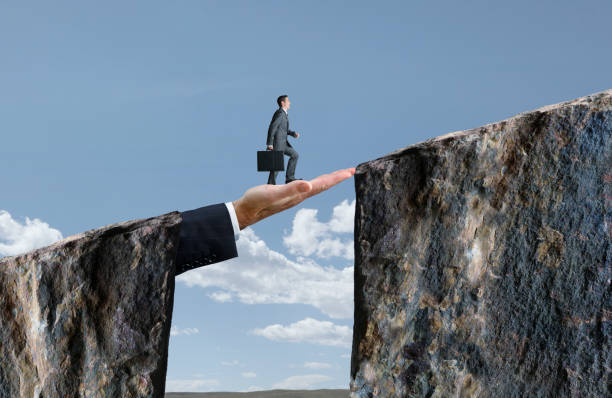Businessman Walking On Hand That Bridges The Gap A businessman holds his briefcase as he walks across a large hand that bridges the gap between two cliffs. bridge hand stock pictures, royalty-free photos & images