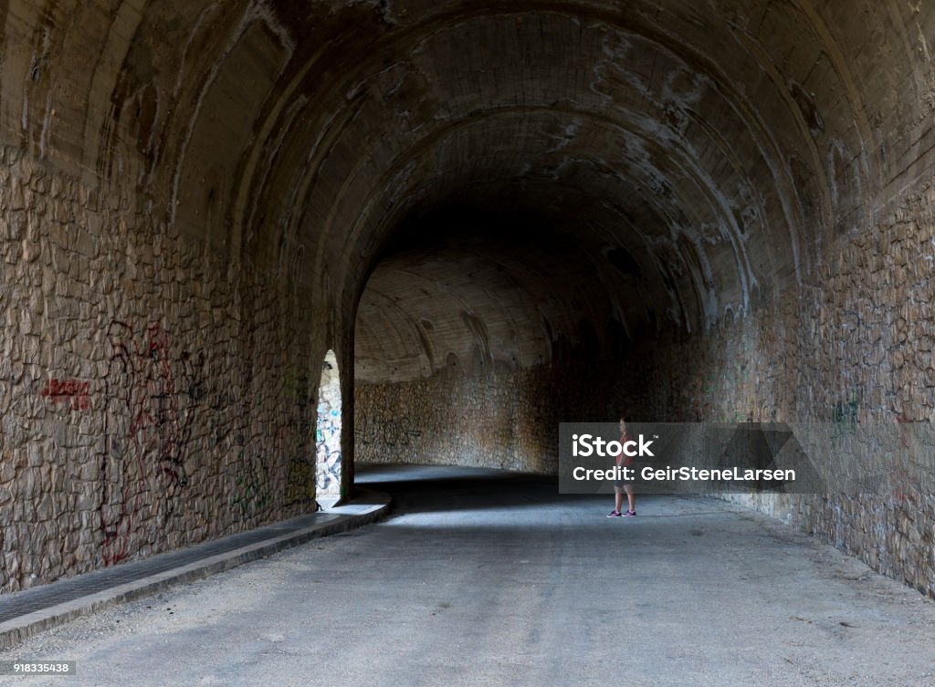 Woman standing in light from an opening in a tunnel Villajoyosa, Spain - March 7, 2017: Woman standing in a tunnel lit up by a beam of light from an opening in the tunnel wall Adult Stock Photo