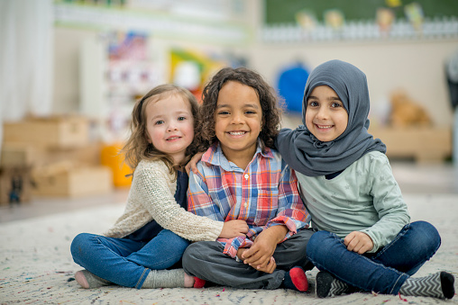 Two girls and a boy are indoors in their preschool classroom. They are smiling at the camera while sitting on the carpet and embracing.