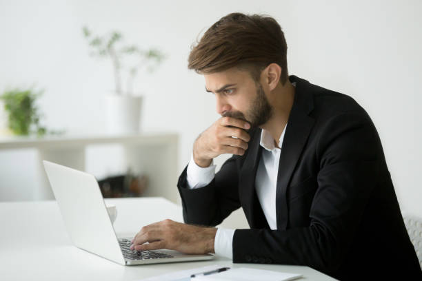 сосредоточенный серьезный бизнесмен мышления чтения онлайн новости с помощью ноутбука - uncertainty men business frowning стоковые фото и изображения