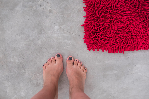 Women feet on a gray concrete floor with red rug fluffy carpet from above. Modern minimal industrial bedroom or bath details