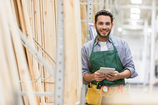 Handsome young Hispanic man works in a lumber yard. He is holding a digital tablet while smiling at the camera.