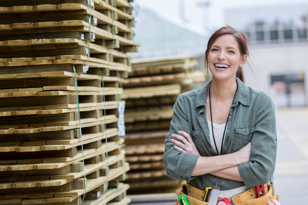 Confident garden center employee Cheerful hardware store employee stands confidently with her arms crossed next to a stack of pallets. woman wearing tool belt stock pictures, royalty-free photos & images