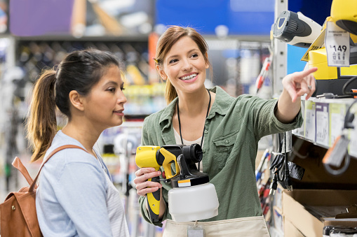 Confident hardware store employee discusses paint sprayers with a female customer. The employee is pointing out different types of sprayers.