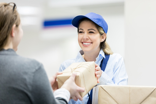 Unrecognizable woman drops off a package at the post office. A female postal worker accepts the package with a smile.