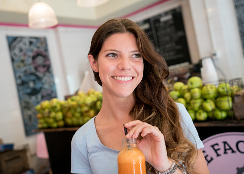 Beautiful female customer enjoying a fresh juice and looking away very happy and smiling at a juice bar