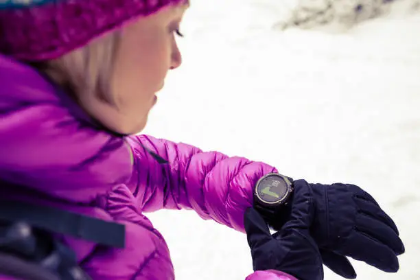 Woman hiker checking the elevation on sports watch, smartwatch with altimeter app in winter woods and mountains. Female trekker in white snowy forest trekking with electronics equipment technology.