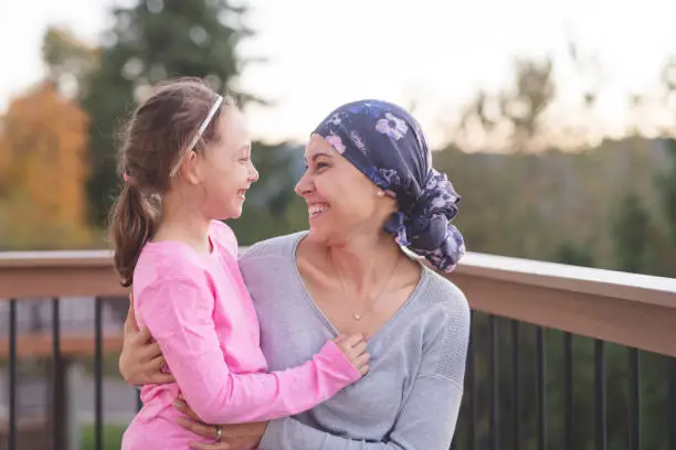 A young mother fighting cancer and wearing a head scarf hugs her daughter and smiles at her deeply as they share a few moments of tranquility together outdoors on a deck.