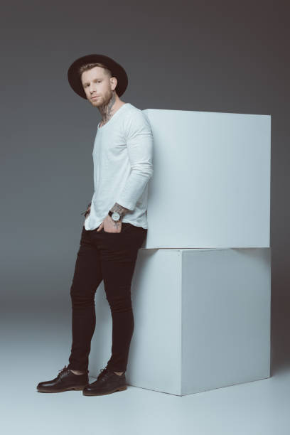 istock stylish young tattooed man in hat standing with hands in pockets and looking at camera on grey 918460600