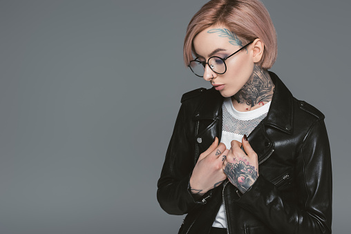 fashionable tattooed girl in eyeglasses and black leather jacket, isolated on grey