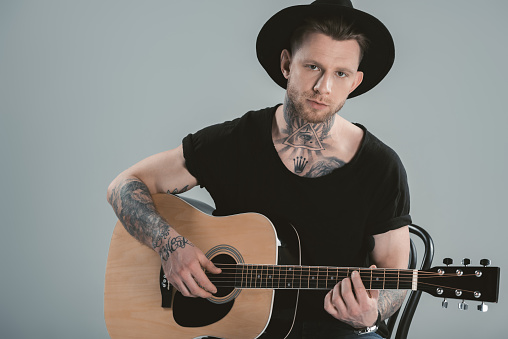 tattooed guitarist in hat playing on acoustic guitar, isolated on grey