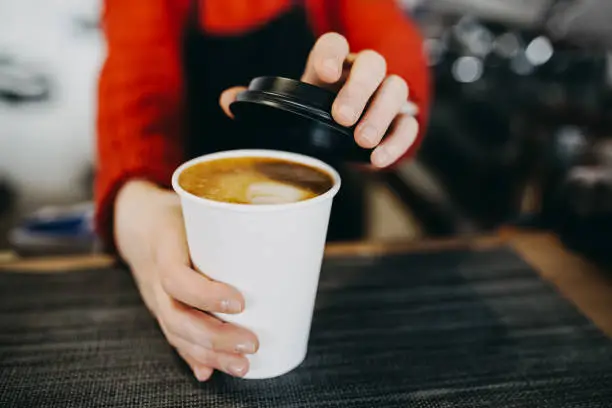 Photo of Barista in apron is holding in hands hot cappuccino in white takeaway paper cup. Coffee take away at cafe shop