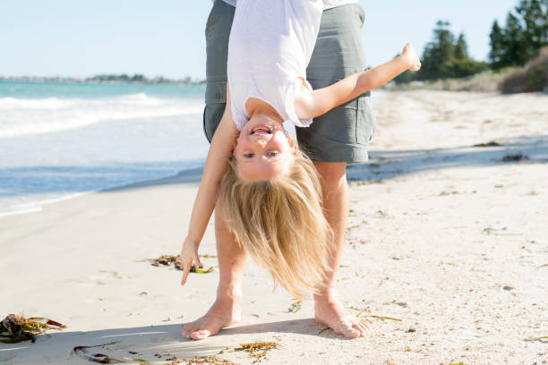father holding sweet young and lovely blond small daughter by her feet playing having fun on the beach in dad and little girl love concept enjoying summer holidays - blond hair carrying little girls small imagens e fotografias de stock