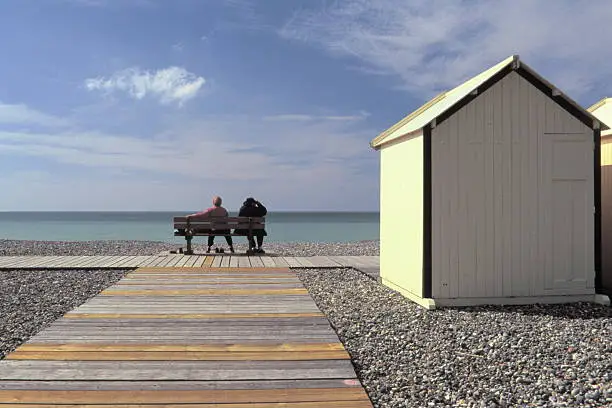 Persons sitting on a bench and looking at the sea besides beach-huts