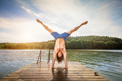 Young man doing a handstand on a wooden jetty at a lake. Summer outdoor activity.