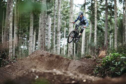 An adult man races down a forest trail, flying through the air from a dirt jump on his mountain bike.  Fun and healthy lifestyle image of recreational outdoor activity.