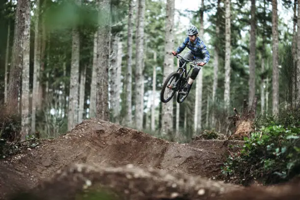 An adult man races down a forest trail, flying through the air from a dirt jump on his mountain bike.  Fun and healthy lifestyle image of recreational outdoor activity.