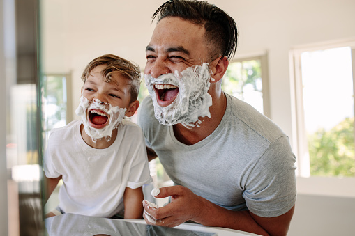 Man and little boy with shaving foam on their faces looking into the bathroom mirror and laughing. Father and son having fun while shaving in bathroom.