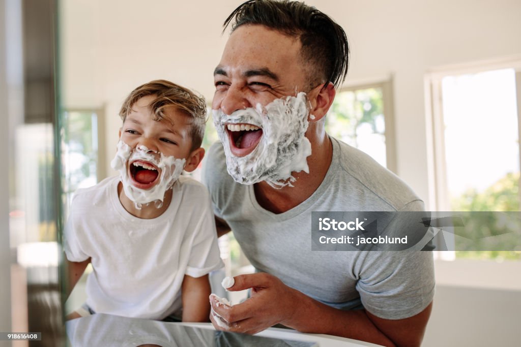 Father and son having fun while shaving in bathroom Man and little boy with shaving foam on their faces looking into the bathroom mirror and laughing. Father and son having fun while shaving in bathroom. Father Stock Photo