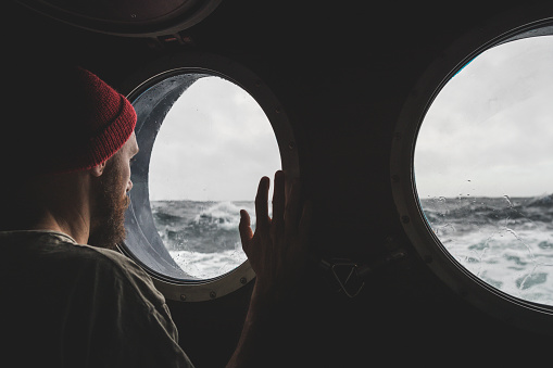 Man at the porthole window of a vessel in a rough sea