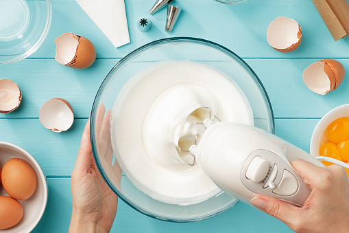 Female hands whipping egg whites cream in glass bowl with mixer on blue wooden table. Step by step recipe of meringue cookies flat lay.