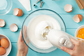 Female hands whipping egg whites cream in glass bowl with mixer on blue wooden table.