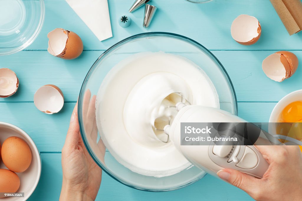Female hands whipping egg whites cream in glass bowl with mixer on blue wooden table. Female hands whipping egg whites cream in glass bowl with mixer on blue wooden table. Step by step recipe of meringue cookies flat lay. Making a Cake Stock Photo