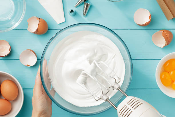 Whipped egg whites cream to perfect peaks in glass bowl with mixer and hand on blue wooden table. stock photo
